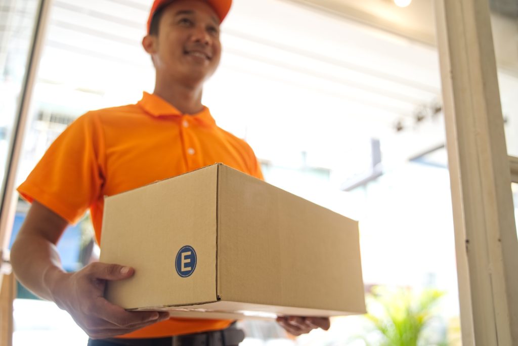 Delivery smiling Asian man in orange uniform and cap holding medium size of post box at the door.